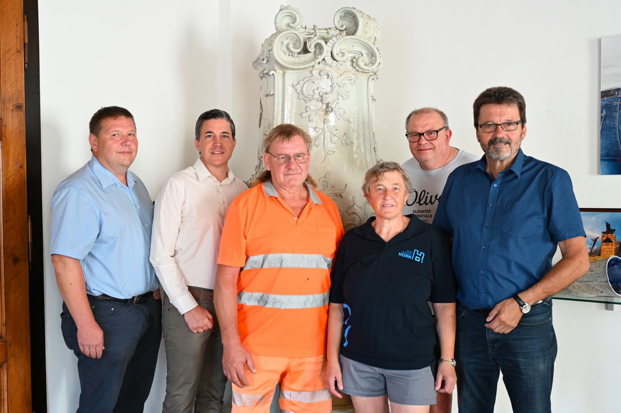 Eine Gruppe von sechs Personen steht vor einem weißen dekorativen Objekt. Der Mann in der Mitte trägt eine orange Sicherheitsuniform. Die Frau rechts trägt ein schwarzes Poloshirt. Der Mann rechts trägt ein blaues Hemd mit Logo.
