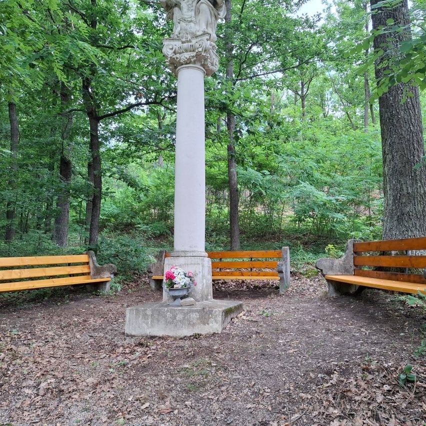 Eine friedliche Parkszene mit einer zentralen Säule, die mit einer Skulptur verziert ist, umgeben von drei Holzbänken und einem Blumenstrauß in einer Vase an der Basis der Säule.