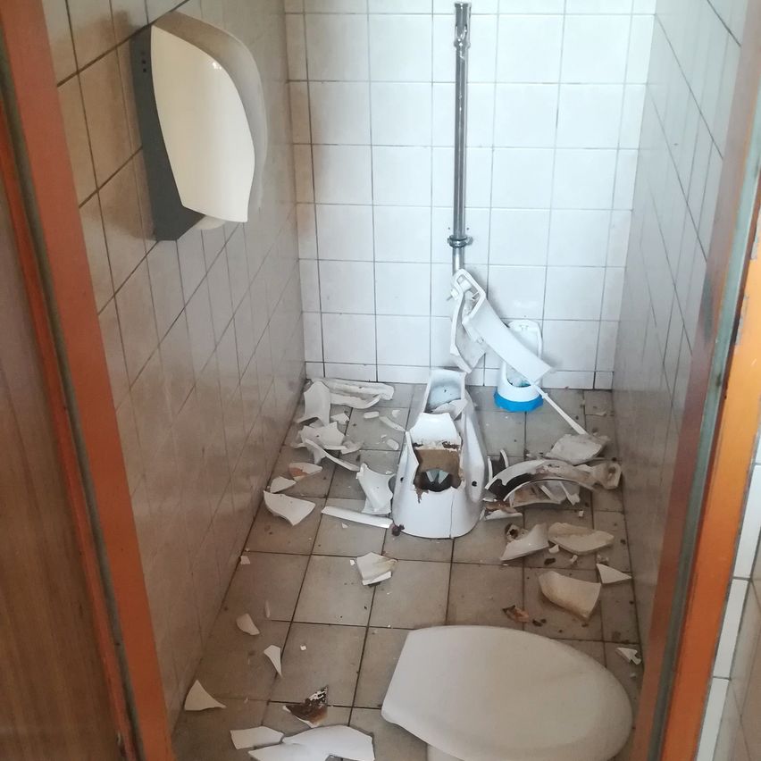 Eine Badezimmerkabine mit einem kaputten Toilettensitz und verstreuten Trümmern auf dem Boden und den Wänden.