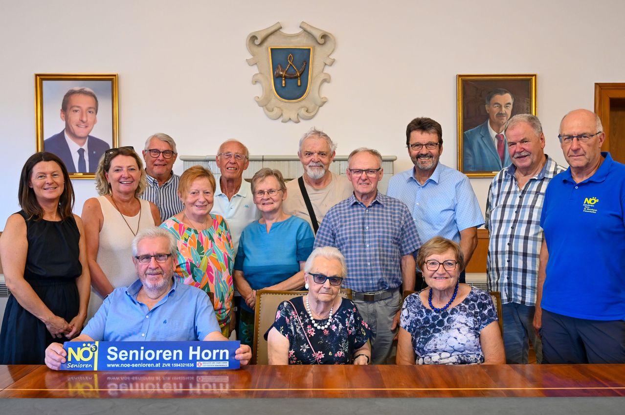 Eine Gruppe älterer Personen, darunter Männer und Frauen, versammelt sich um einen Tisch mit einem blauen Schild, auf dem 'Senioren Horn' steht. Sie alle lächeln und posieren für ein Foto. Hinter ihnen befindet sich eine Wand mit einem gerahmten Bild und einem dekorativen Schild.