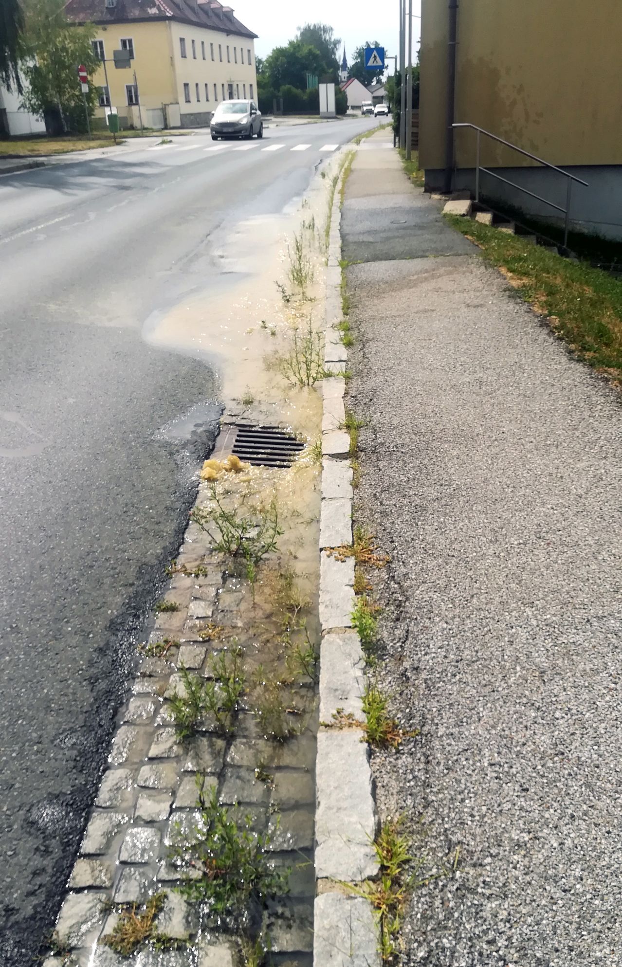 A street with a flooded curb and a drain, next to a gravel pathway.