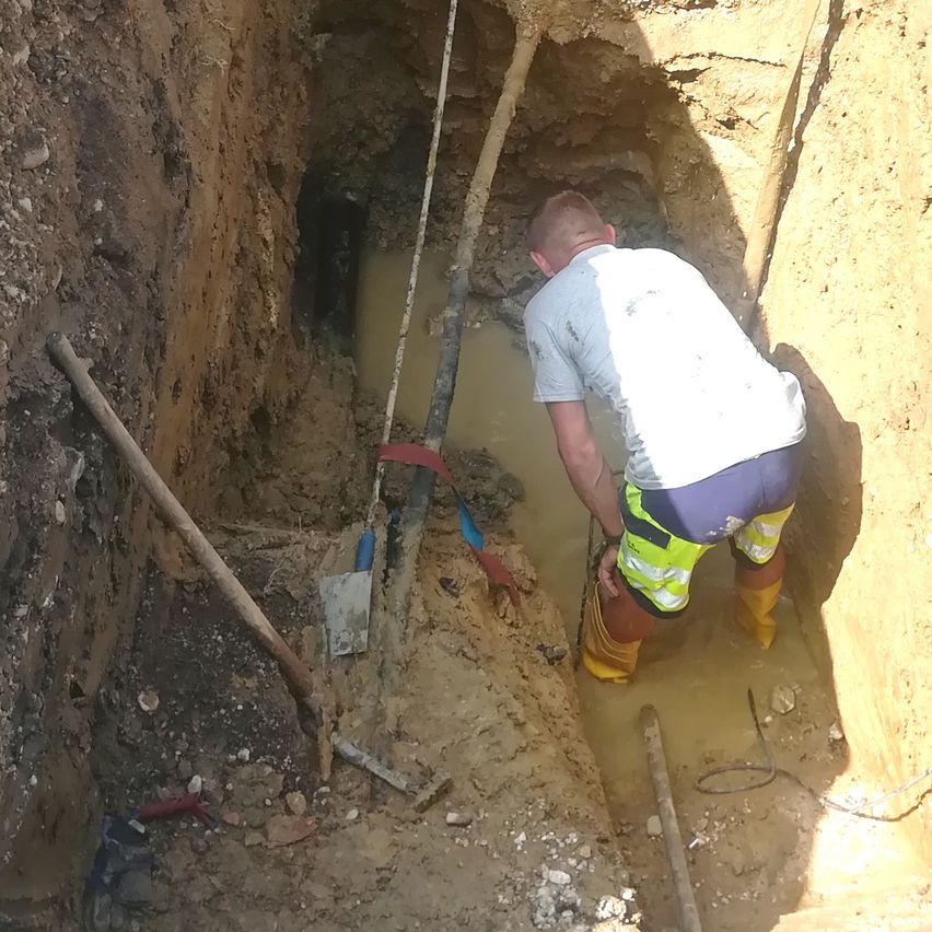 A man in a white shirt and yellow boots is working in a muddy pit, possibly installing a pipe. Tools and a shovel are nearby.