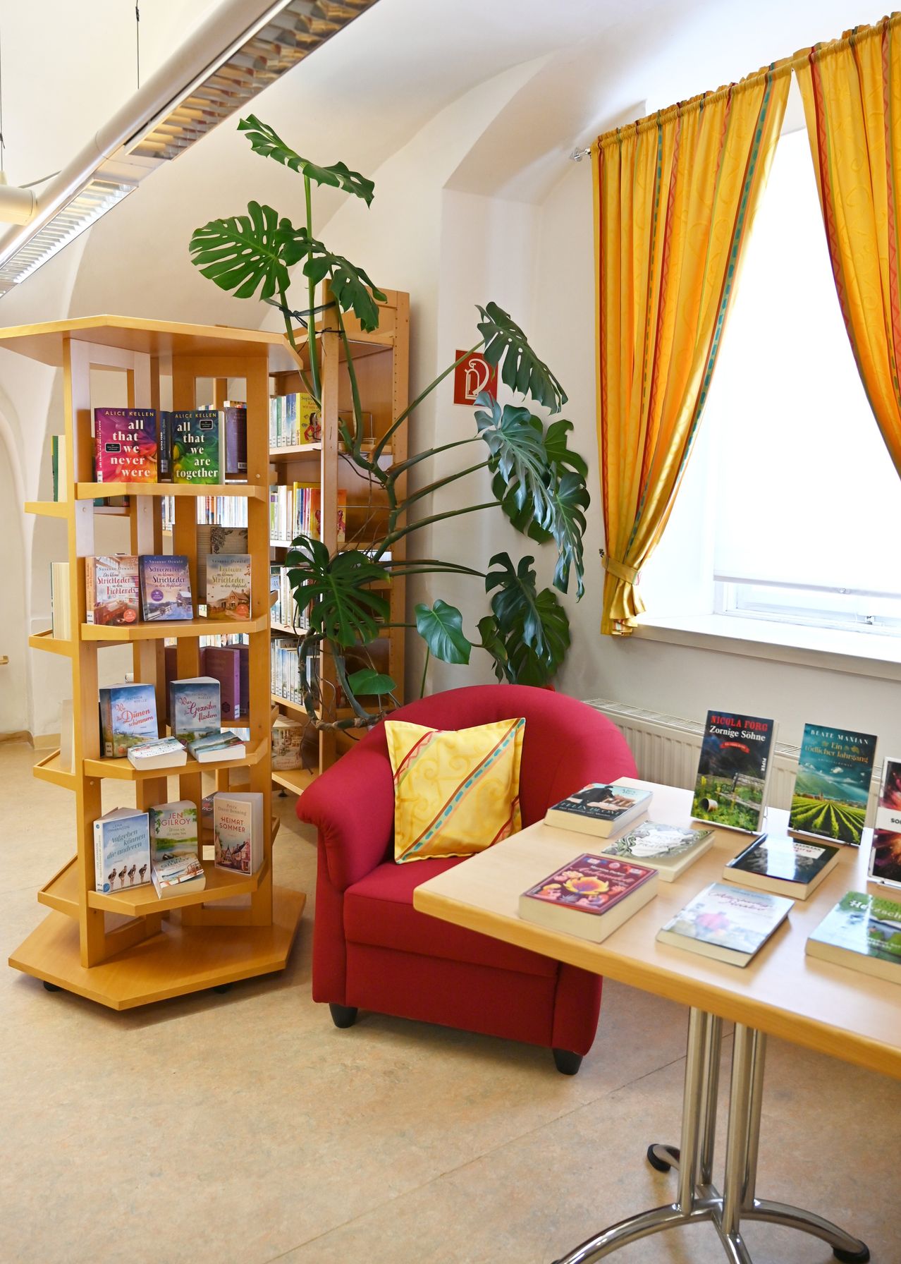 A cozy reading nook with a red chair, a wooden table with books, and a bookshelf filled with books. A large green plant is in the background.