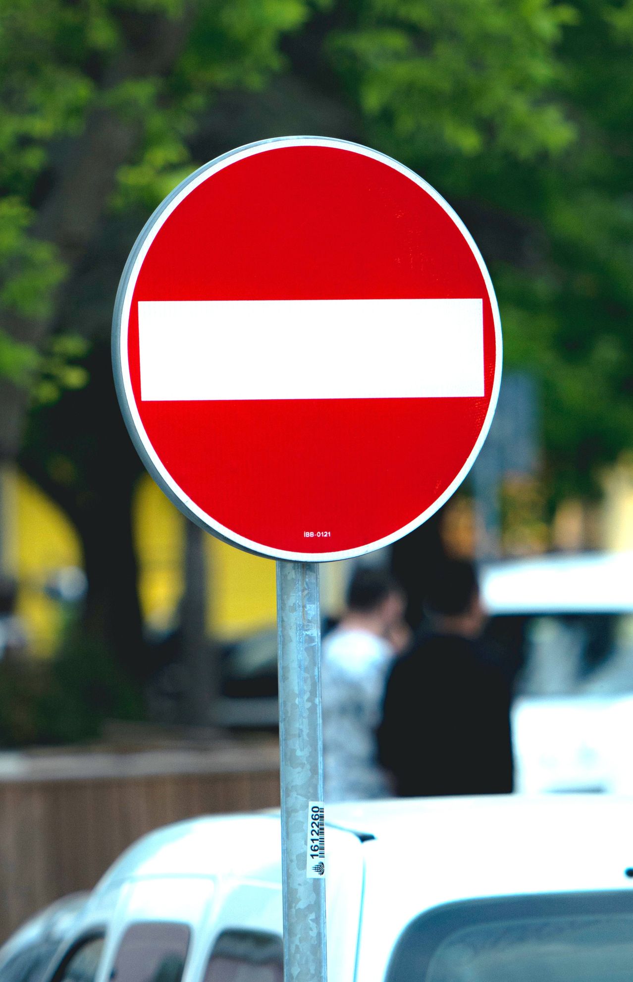 A red circular sign with a white bar through the center, indicating a prohibition or restriction. The background is blurred with people and a vehicle.