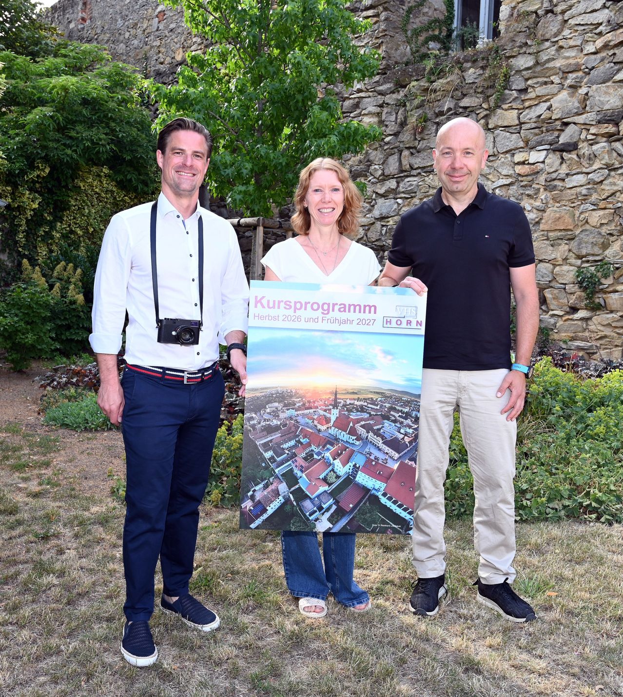 Three people stand in front of a stone wall, holding a poster with a city skyline and the text 'Kursprogramm Herbst 2026 und Frühjahr 2027'. The man on the left wears a white shirt and blue pants, the woman in the middle wears a white top and blue jeans, and the man on the right wears a black shirt and khaki pants.