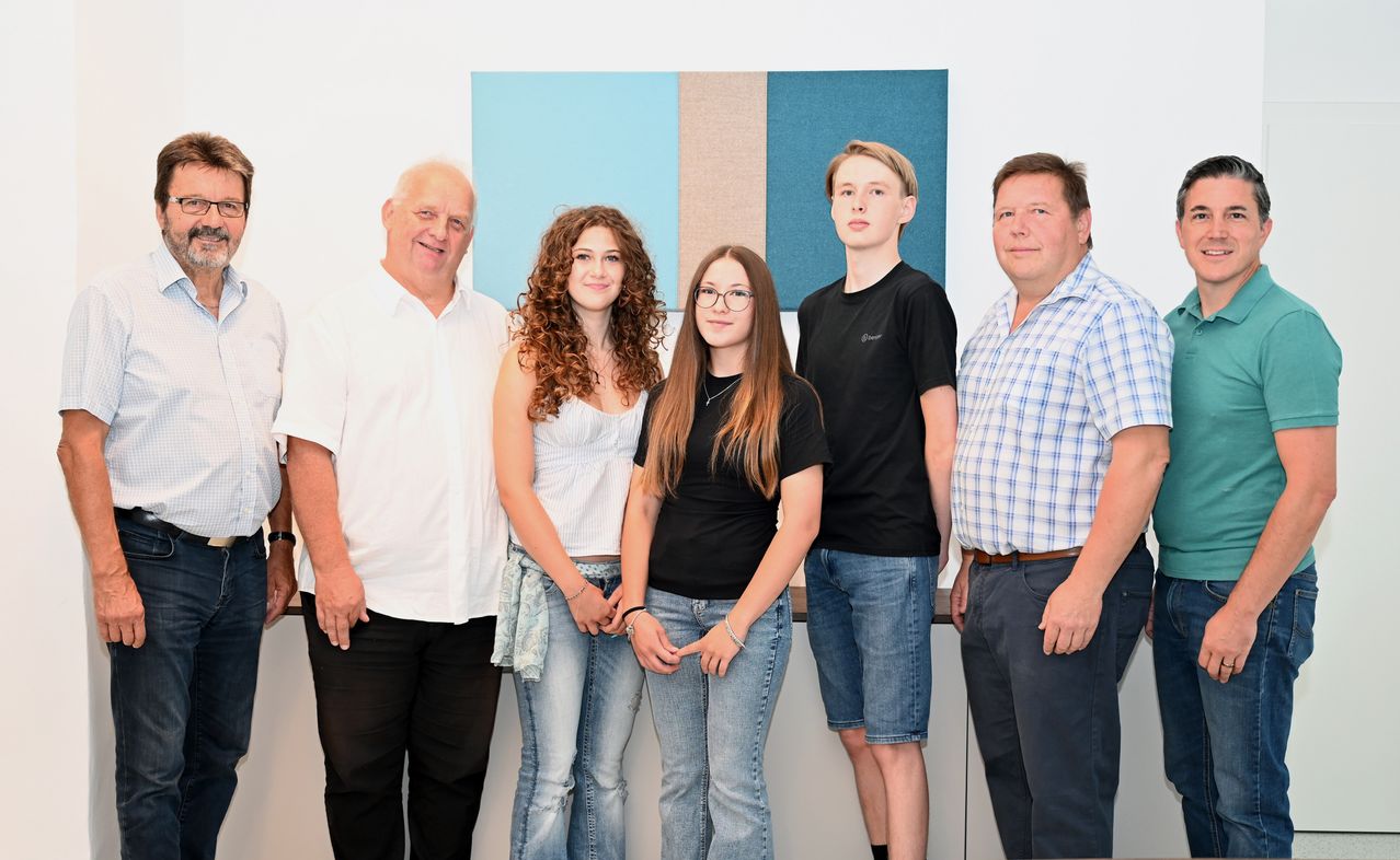 Five people are standing together in front of a white wall, smiling for a photo. The two women are wearing glasses and the two men are wearing belts.