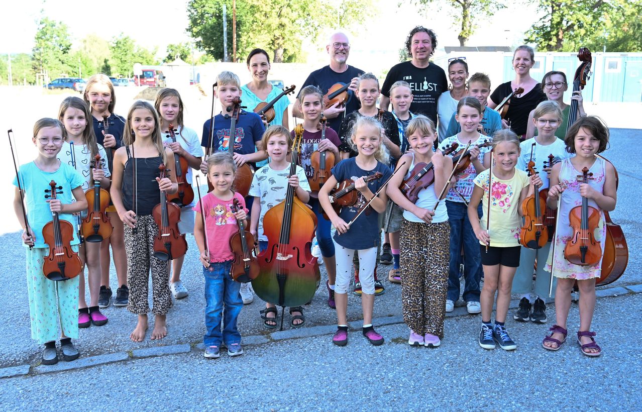 A group of children and adults are posing for a photo while holding violins and cellos. They are standing on a gravel surface.