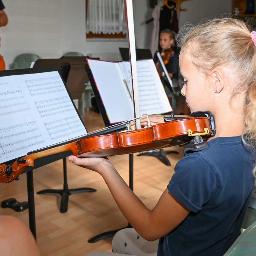 A young girl in a blue shirt practices playing the violin in a music room with sheet music on stands.