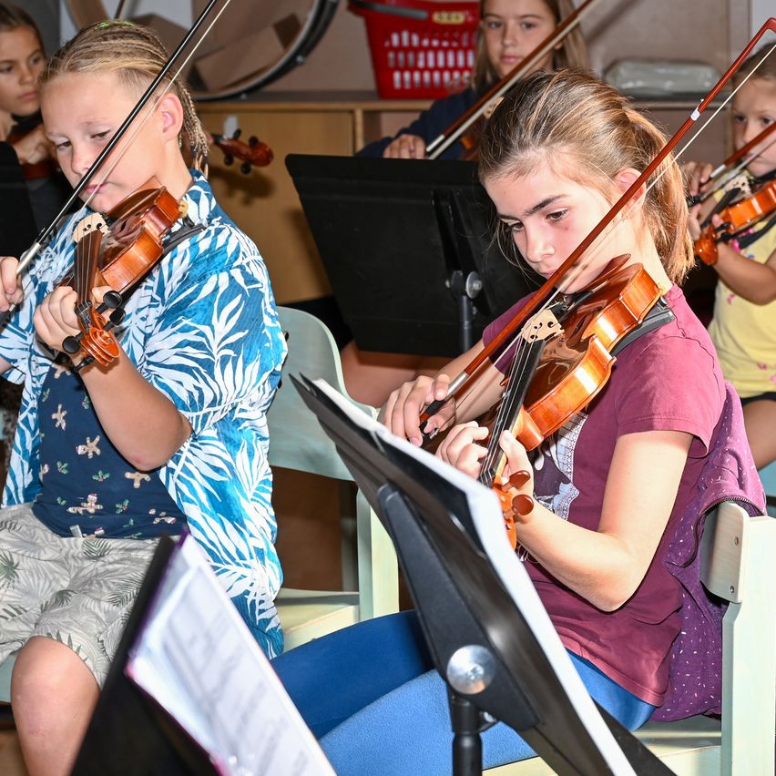 A group of young girls are playing violins in a classroom setting. They are seated and focused on their music sheets.