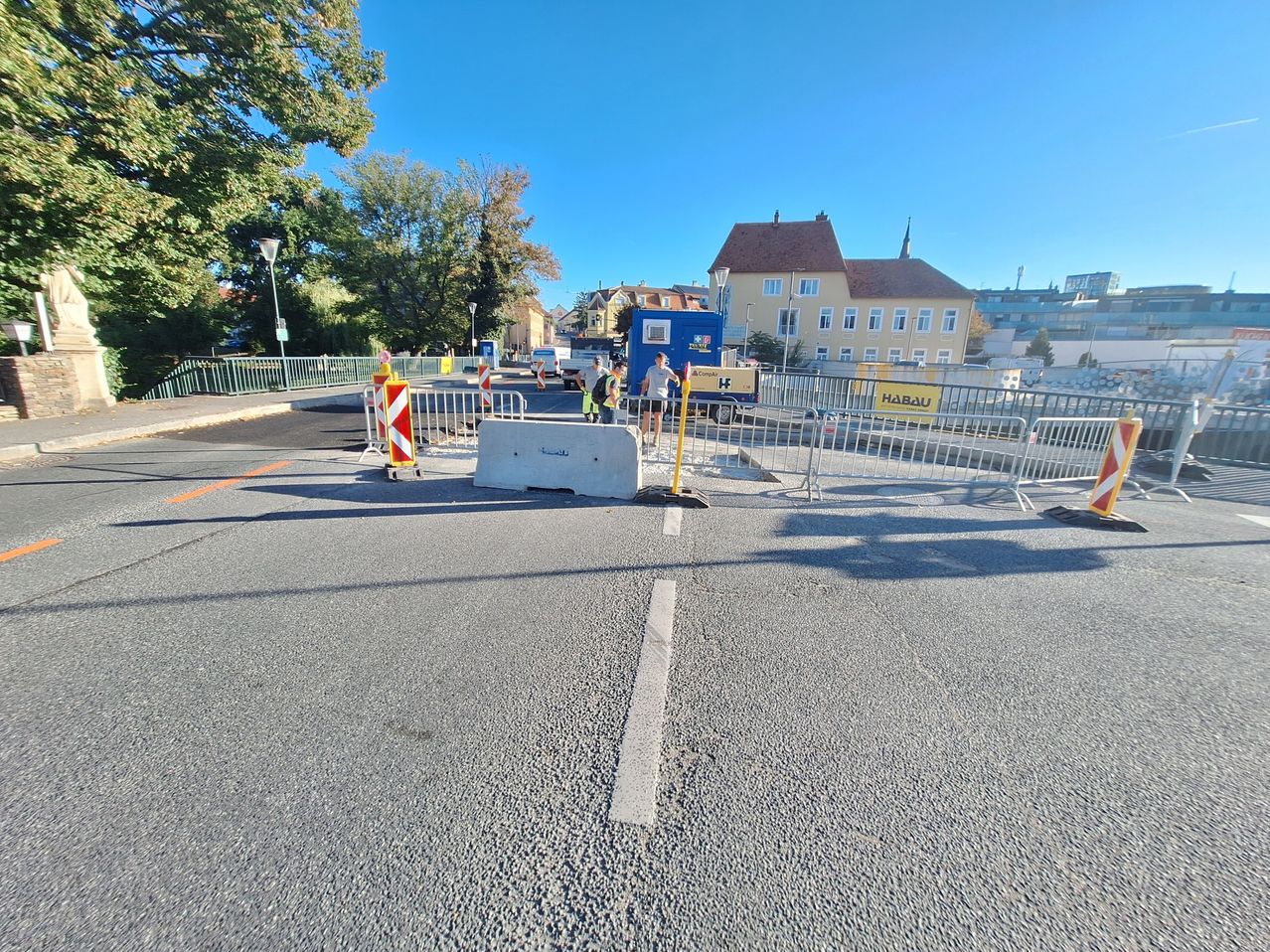 A road construction site with workers and barriers, a blue truck, and a yellow sign. Trees and buildings are in the background.