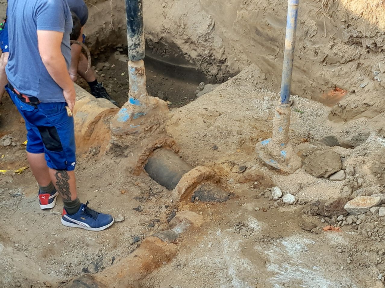 Two men are digging a hole in the ground with a large pipe in the middle. One man is standing and holding a tool.