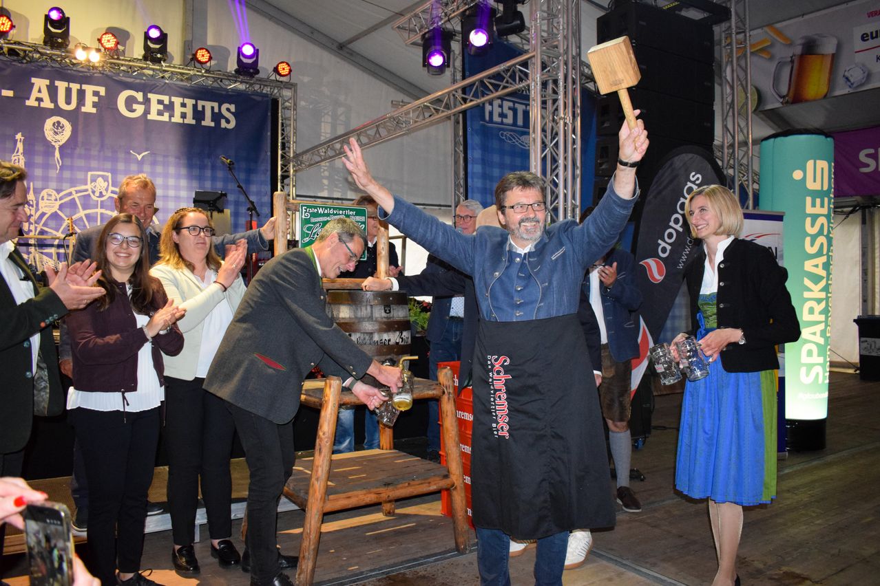 Ein Mann mit einem Hammer steht auf einer Bühne mit einem anderen Mann, der Bier einschenkt. Ein Fass und ein Tisch sind hinter ihnen. Zuschauer beobachten, einschließlich einer Frau, die ein Glas hält.