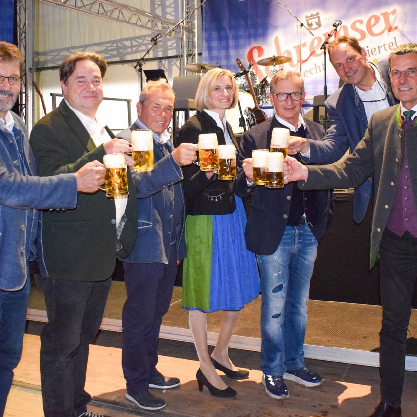 Sieben Männer und eine Frau, gekleidet in formeller Kleidung, sind auf der Bühne, lächeln und halten Gläser mit Bier hoch. Hinter ihnen ist ein Banner mit dem Text 'Schlenser' und Musikinstrumente auf der Bühne.