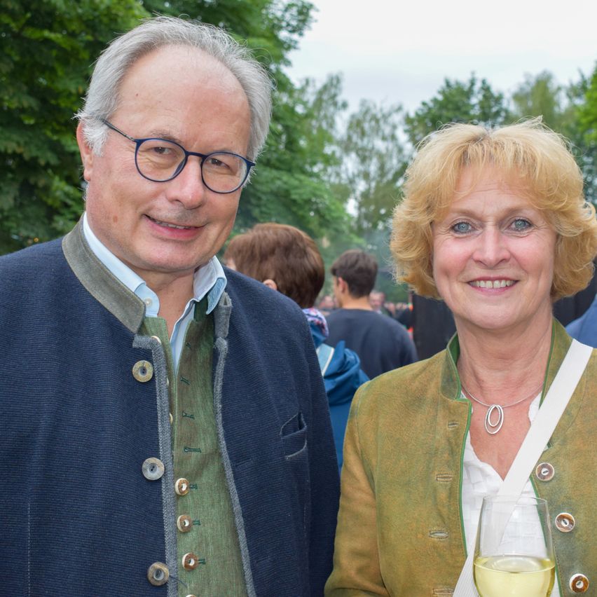 Ein Mann und eine Frau stehen lächelnd in einem Park mit Bäumen und Menschen im Hintergrund. Der Mann trägt eine Brille und einen blauen Mantel, während die Frau ein Weinglas hält.
