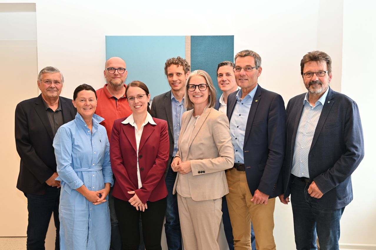 Eine Gruppe von Fachleuten in Geschäftskleidung steht in einem Raum und lächelt für ein Foto. Der Hintergrund ist schlicht und die Beleuchtung ist hell.
