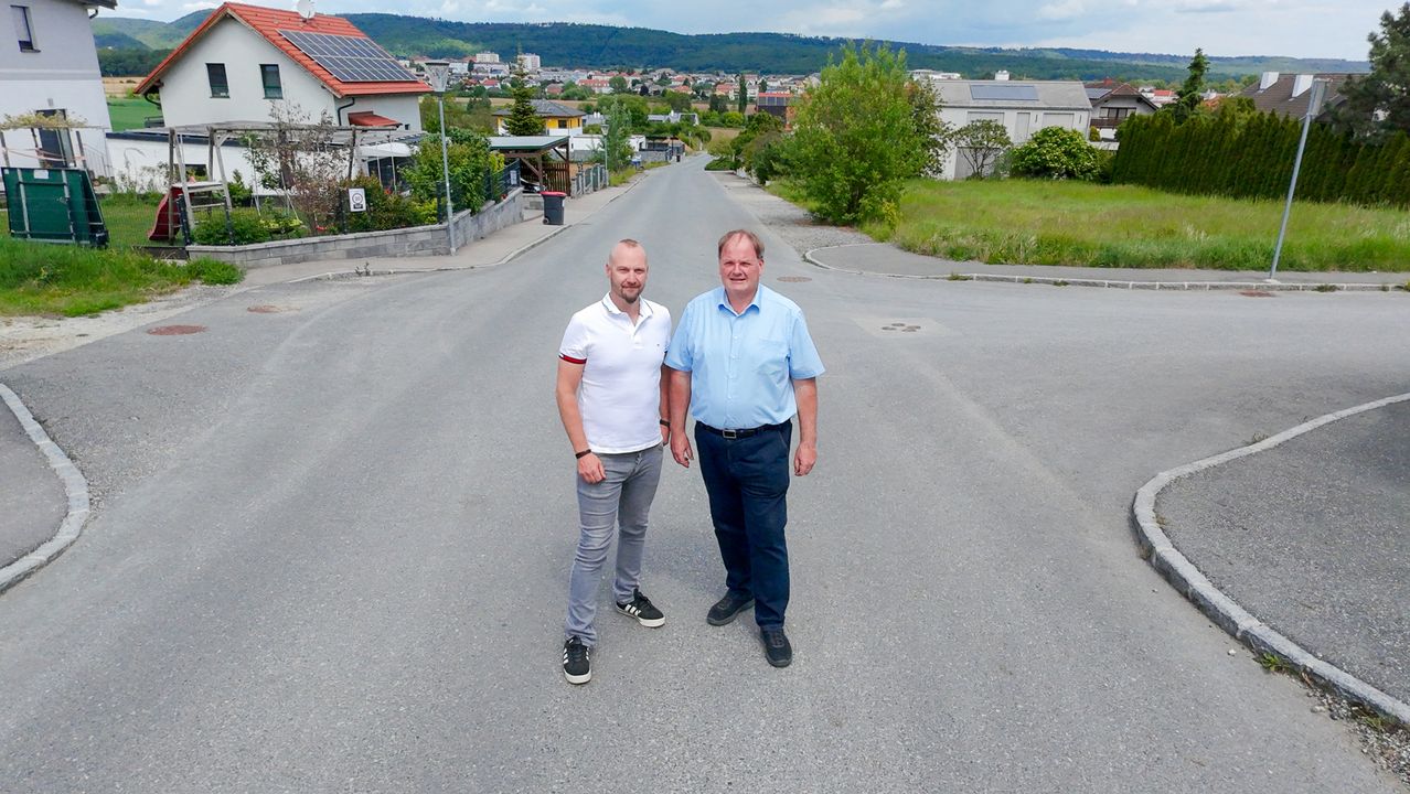 Zwei Männer stehen in der Mitte einer Straße mit Häusern und Grünflächen auf beiden Seiten. Sie lächeln, und die Straße ist leer. Im Hintergrund befindet sich eine Stadtlandschaft mit Gebäuden und Bergen.