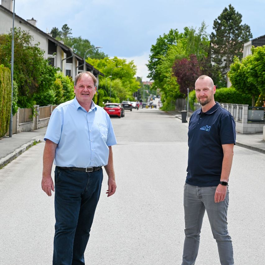 Zwei Männer stehen in der Mitte einer Straße und lächeln für ein Foto. Bäume und Gebäude sind im Hintergrund zu sehen.