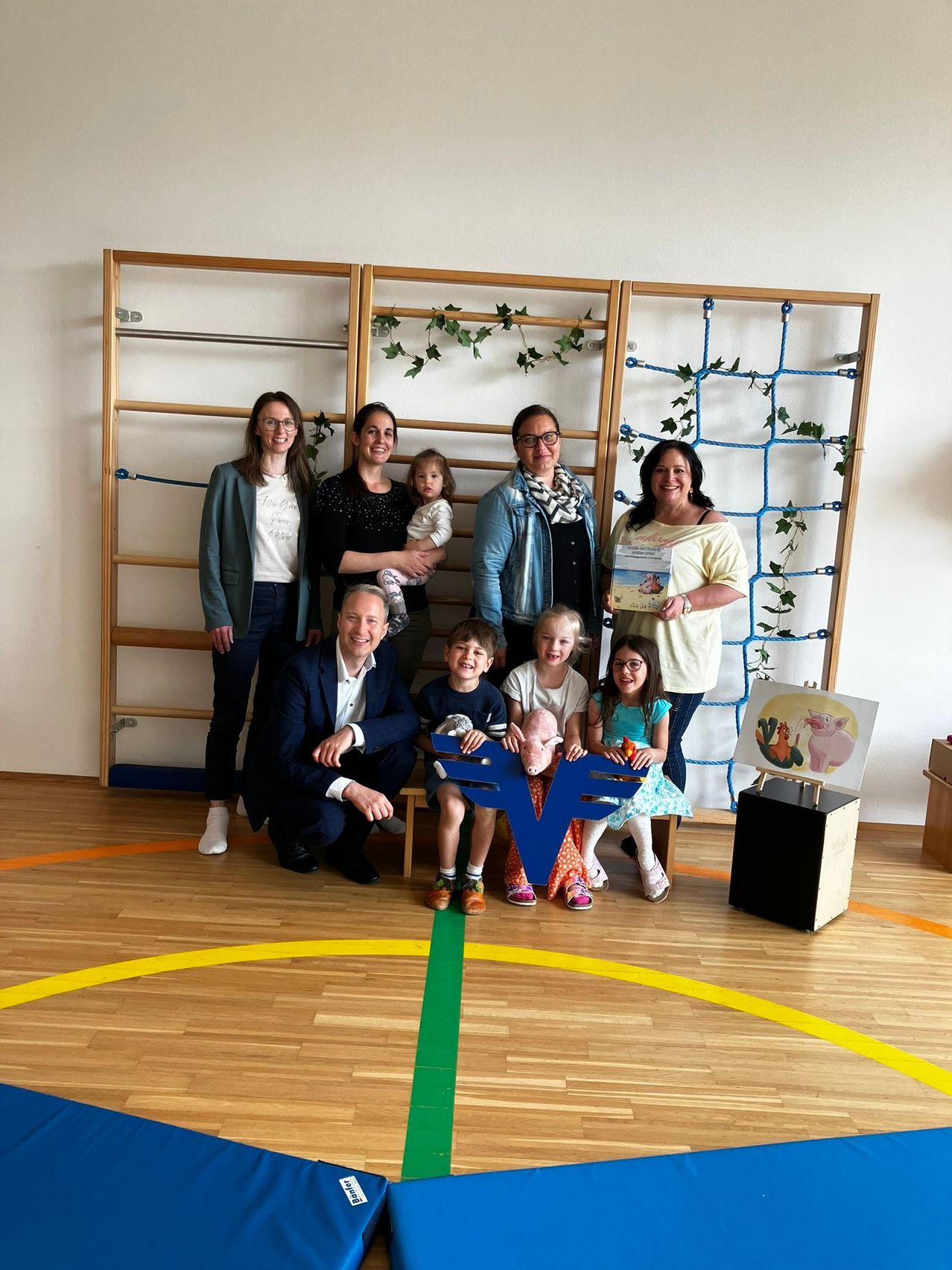 Eine Gruppe von Erwachsenen und Kindern in einem Klassenzimmer mit blauer Wand, Holzboden und Kletterwand. Ein Mann hält einen blauen Buchstaben E und eine Frau ein Zertifikat.