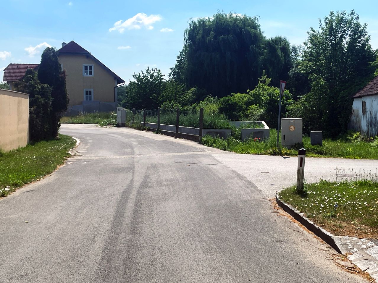 Eine ländliche Straße mit einem Haus auf der linken Seite und Grünflächen entlang der Straßenseiten. Bäume und Büsche befinden sich auf der rechten Seite. Der Himmel ist klar mit einigen Wolken.
