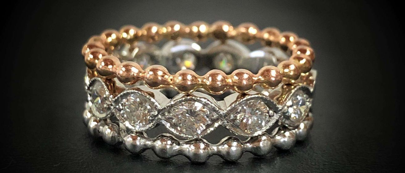 Drei gestapelte Ringe mit Roségold-, Weißgold- und Silberbändern, verziert mit Diamanten, Perlen und filigranen Mustern auf einer schwarzen Oberfläche.