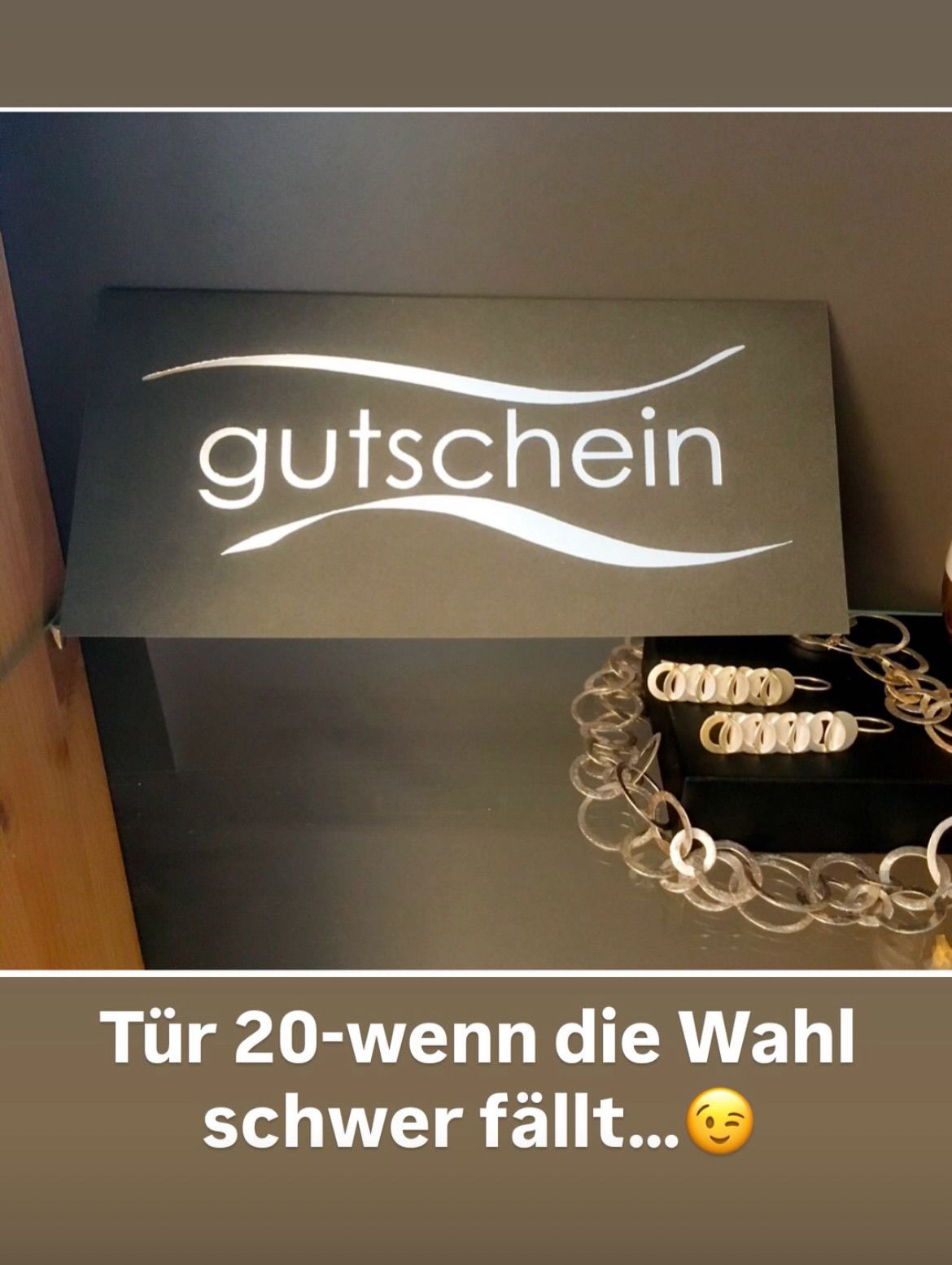 Ein Schild mit dem Wort 'gutschein' über einer Schmuck-Ausstellung. Der Hintergrund ist grau und eine schwarze Box mit einem goldenen Kettendesign steht auf einer Glasfläche. Der Text unter dem Bild lautet 'Tür 20-wenn die Wahl'.