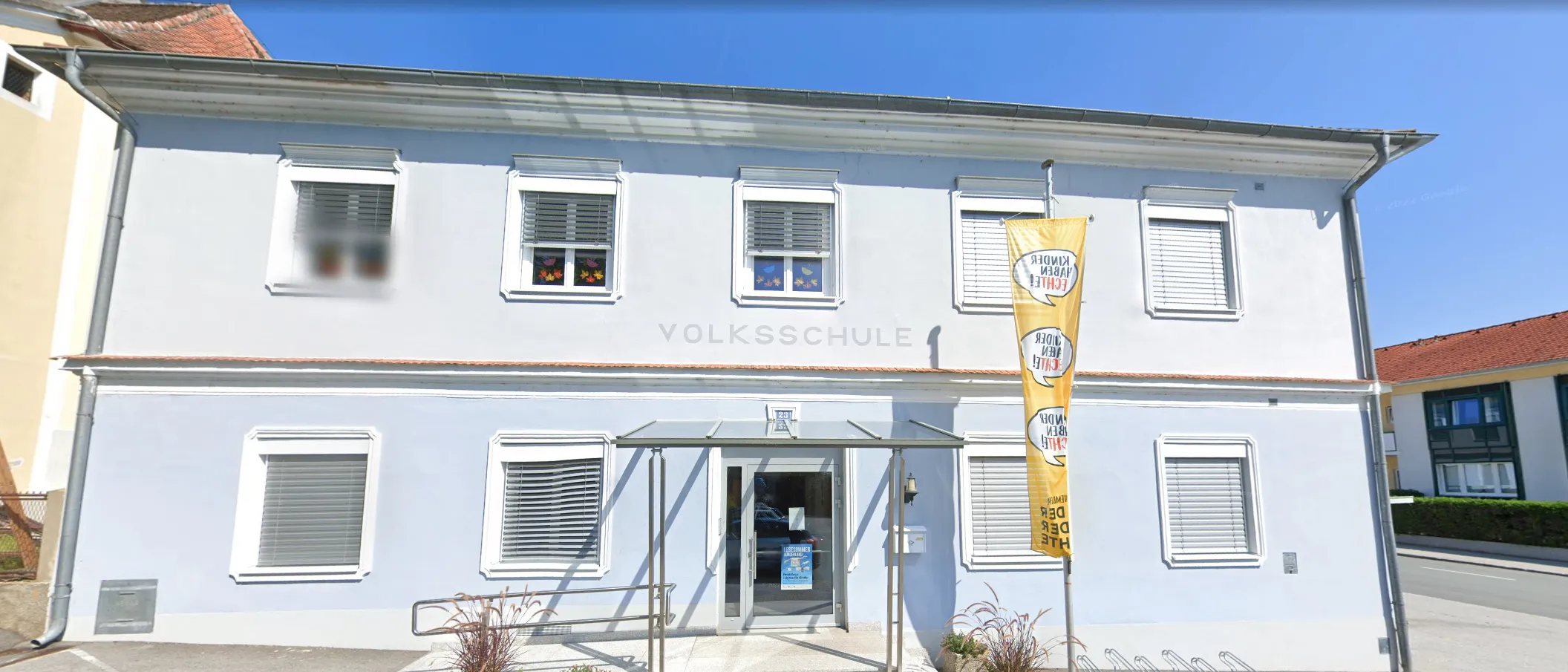 Ein hellblaues Gebäude mit weißen Fensterläden und einem Glasvordach über dem Eingang. Das Gebäude trägt oben 'Volksschule'. Eine gelbe Fahne hängt an der rechten Seite.