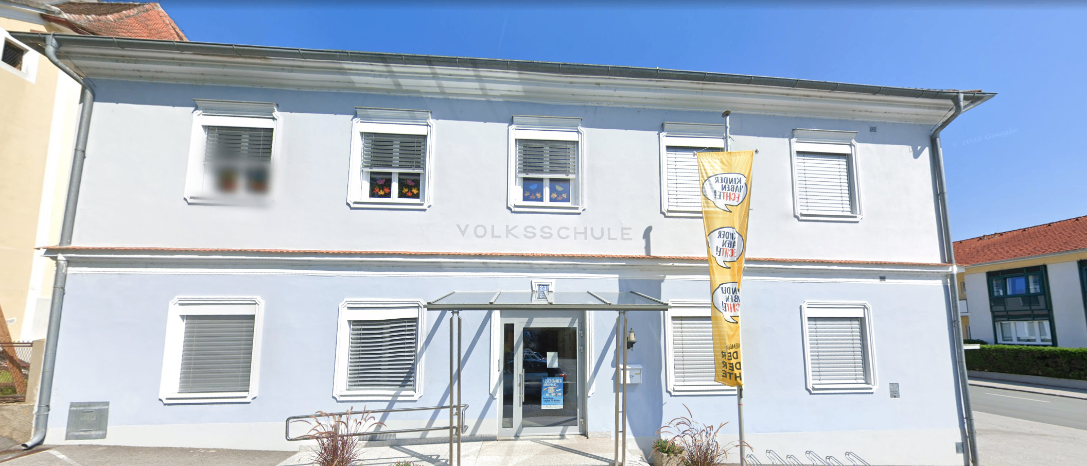 Ein hellblaues Gebäude mit weißen Fensterläden und einem Glasvordach über dem Eingang. Das Gebäude trägt oben 'Volksschule'. Eine gelbe Fahne hängt an der rechten Seite.