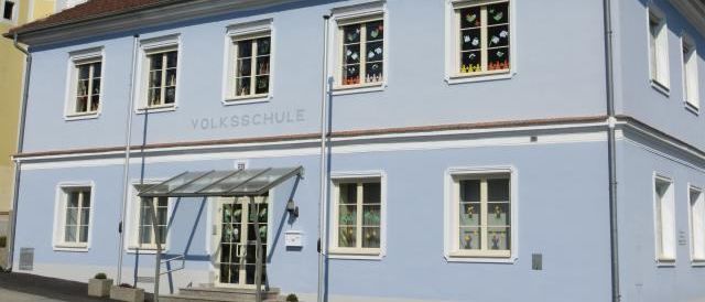 Ein hellblaues Gebäude mit braunem Dach und weißem Rand hat das Wort Volksschule über dem Eingang. Über dem Eingang befindet sich ein Vordach mit mehreren Stufen, die hinaufführen.