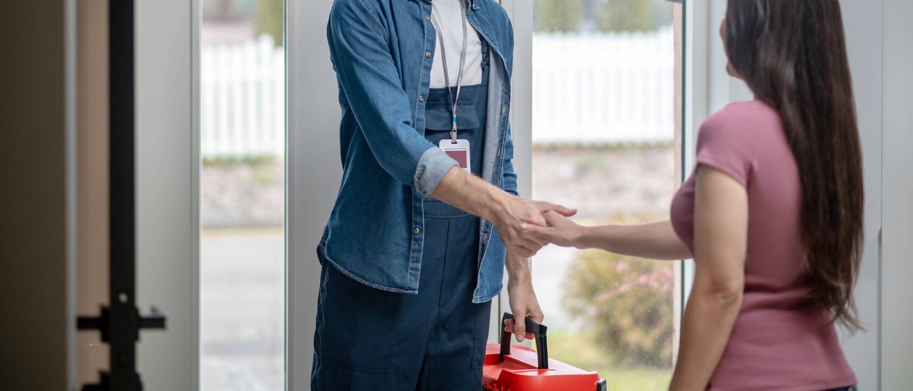 Ein lächelnder Mann in Arbeitskleidung gibt einer Frau an einer Glastür die Hand und hält eine rote Werkzeugkiste.
