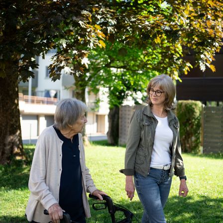 Zwei Frauen gehen in einem Park, eine benutzt einen Gehstock. Sie stehen unter einem Baum mit einem Gebäude und einem Zaun im Hintergrund.