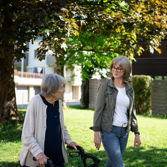 Zwei Frauen gehen in einem Park, eine benutzt einen Gehstock. Sie stehen unter einem Baum mit einem Gebäude und einem Zaun im Hintergrund.