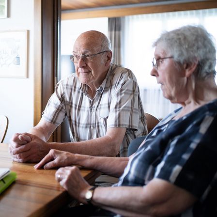 Ein älterer Mann mit Brille sitzt an einem Holztisch mit einer älteren Frau und führt ein Gespräch. Dahinter ziert ein gerahmtes Bild die Wand, und ein Stuhl mit einem Buch steht in der Nähe.