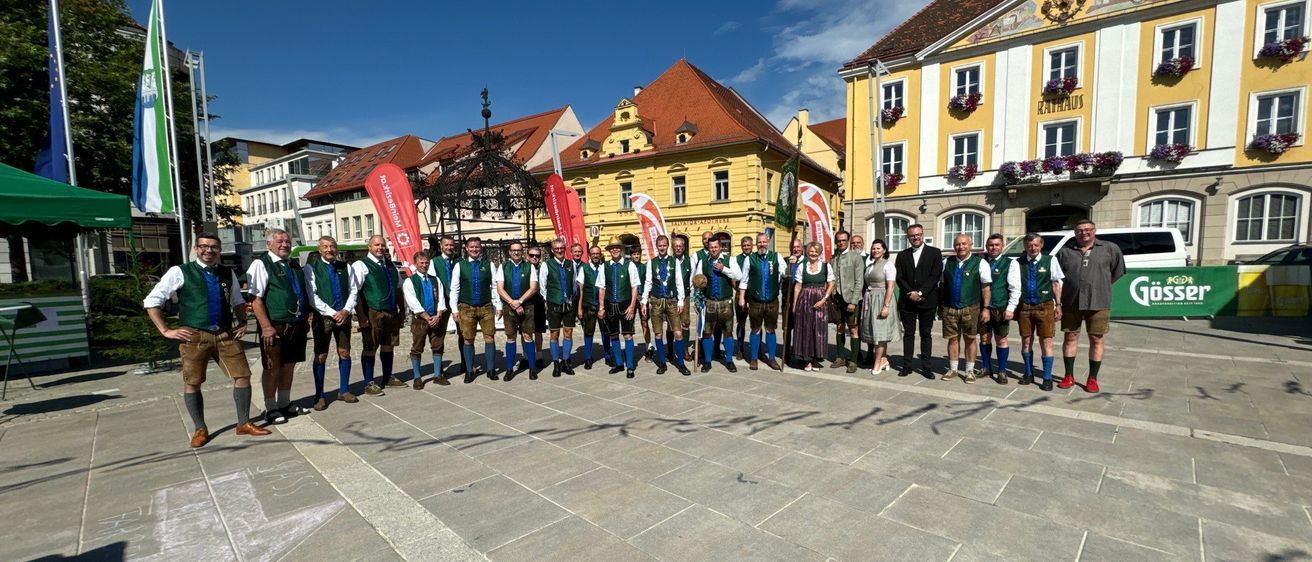 Eine Gruppe von Menschen in traditioneller bayerischer Kleidung posiert für ein Foto auf einem Stadtplatz. Hinter ihnen stehen Gebäude mit gelben Fassaden und roten Dächern.