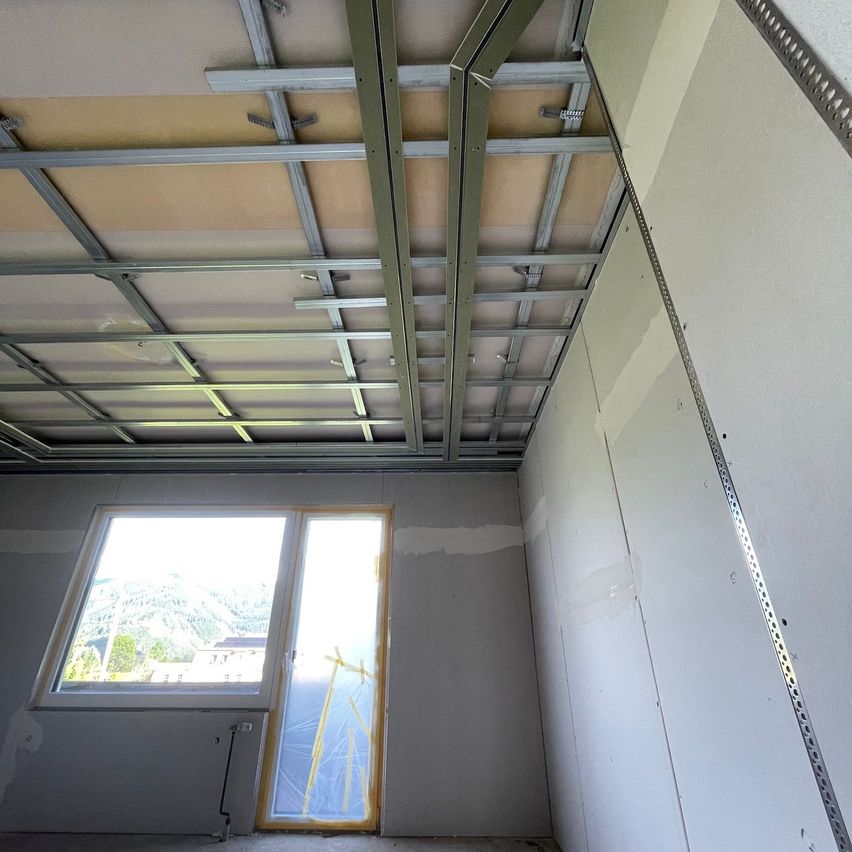 An interior room with exposed steel beams on the ceiling and drywall on the walls. A window is partially visible.