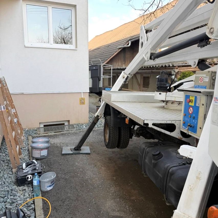 Ein weißer Lastwagen mit ausgestrecktem Arm ist vor einem Haus mit Fenster und Dach geparkt. Verschiedene Gegenstände, darunter Eimer und eine Flasche, liegen auf dem Boden.