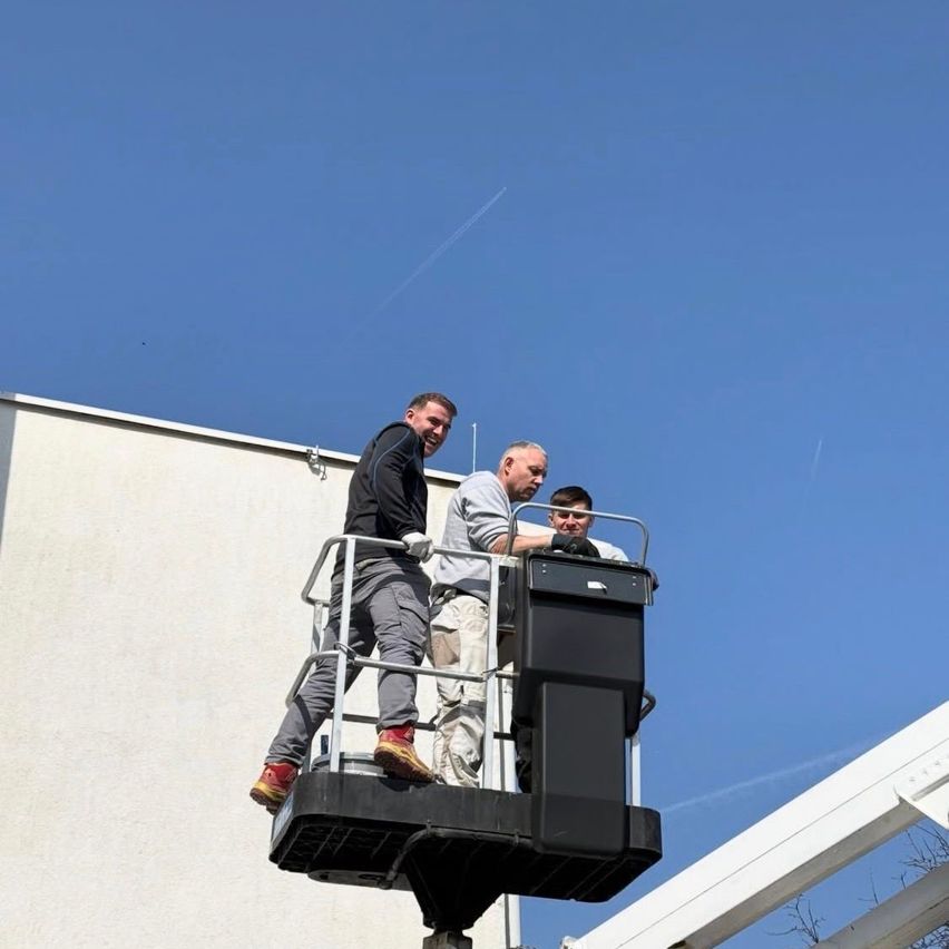 Drei Männer sind auf einer Plattform und arbeiten an einem Gebäude. Ein Mann lächelt, ein anderer schaut nach unten und der dritte ist dazwischen. Die Plattform ist erhöht und das Gebäude hat eine weiße Wand.