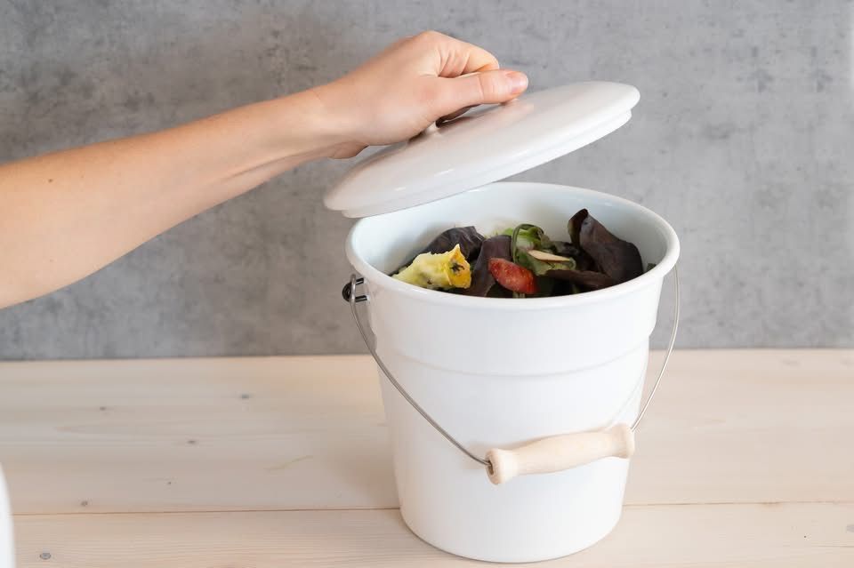 Eine Person öffnet einen weißen Kompostbehälter, der mit Gemüseresten und Fruchtschalen auf einem Holztisch gefüllt ist.