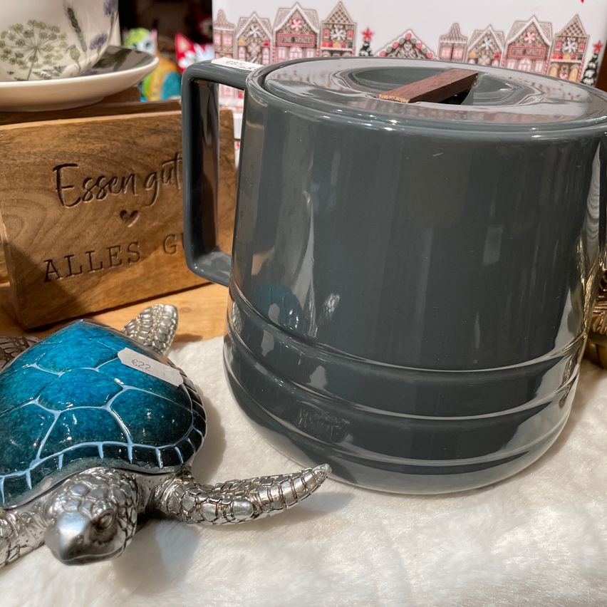 Ein grauer Keramikkessel mit einem Holzdeckel steht neben einer silberfarbenen Schildkrötenfigur auf einer weißen Oberfläche. Dahinter befindet sich ein Holzkasten mit der Aufschrift 'Essen gut'.