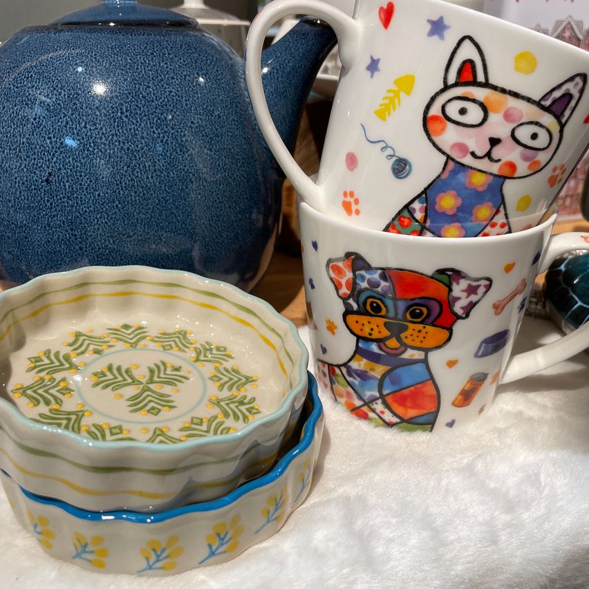 Zwei Tassen mit bunten Katzen- und Hundedesigns, gestapelt auf einem kleinen, gemusterten Teller, neben einem blauen Teepott auf einem weißen Tuch.