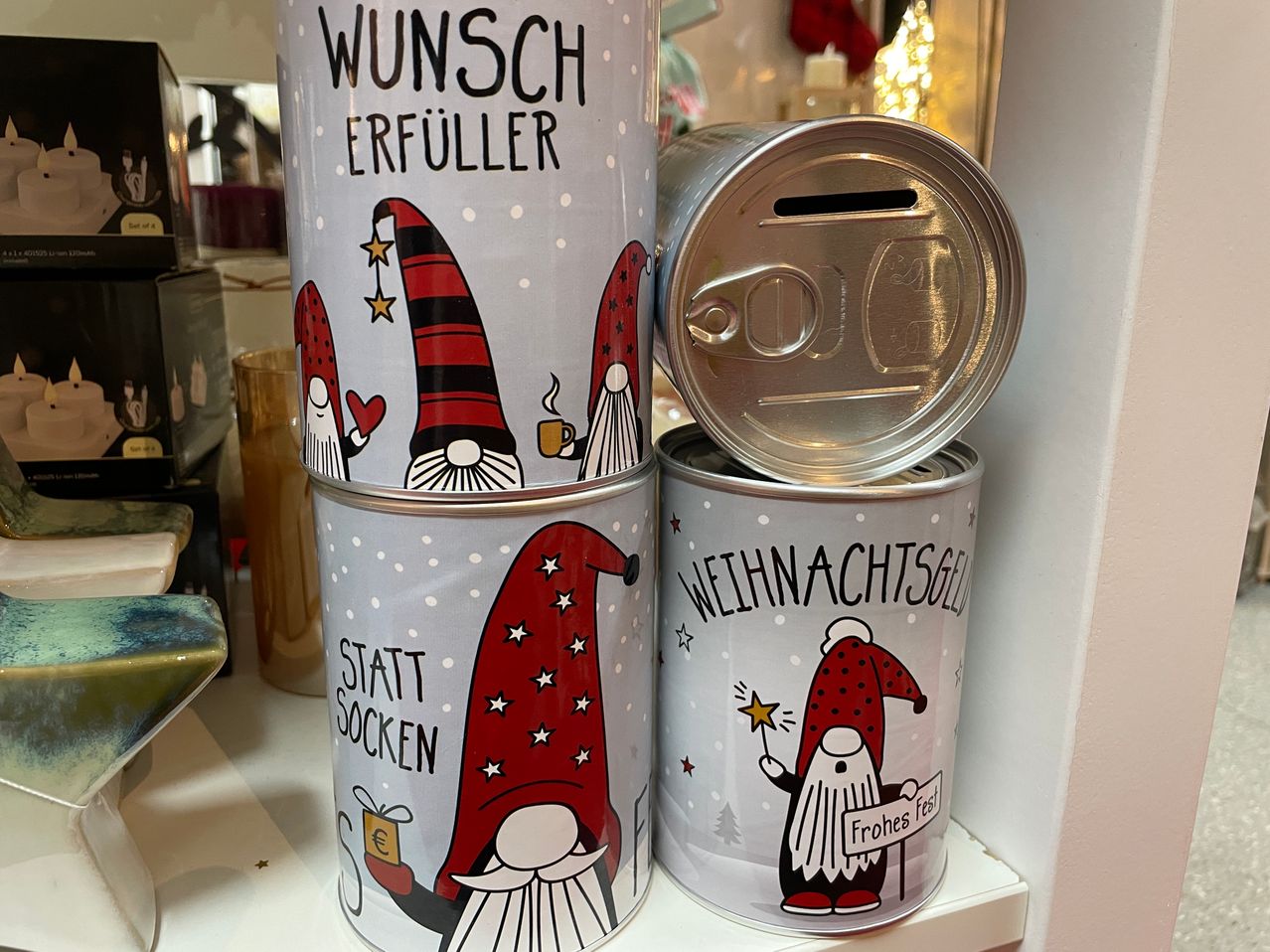 Drei silberne Blechdosen mit Weihnachtsdekorationen sind auf einem Regal gestapelt. Jede Dose hat ein anderes Design: Die oberste sagt 'Wunsch Erfüller', die mittlere sagt 'Statt Socken' und die unterste sagt 'Weihnachtsgeld'.