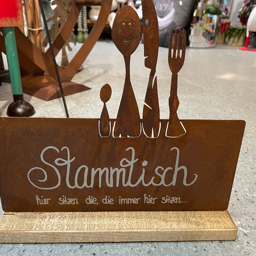 Ein rostige Metallschild mit einer Familie von Küchenutensilien über dem Wort Stammtisch, was Familientisch auf Deutsch bedeutet. Die Utensilien sind Gabel, Messer, Löffel und ein Baby. Der Hintergrund ist verschwommen.
