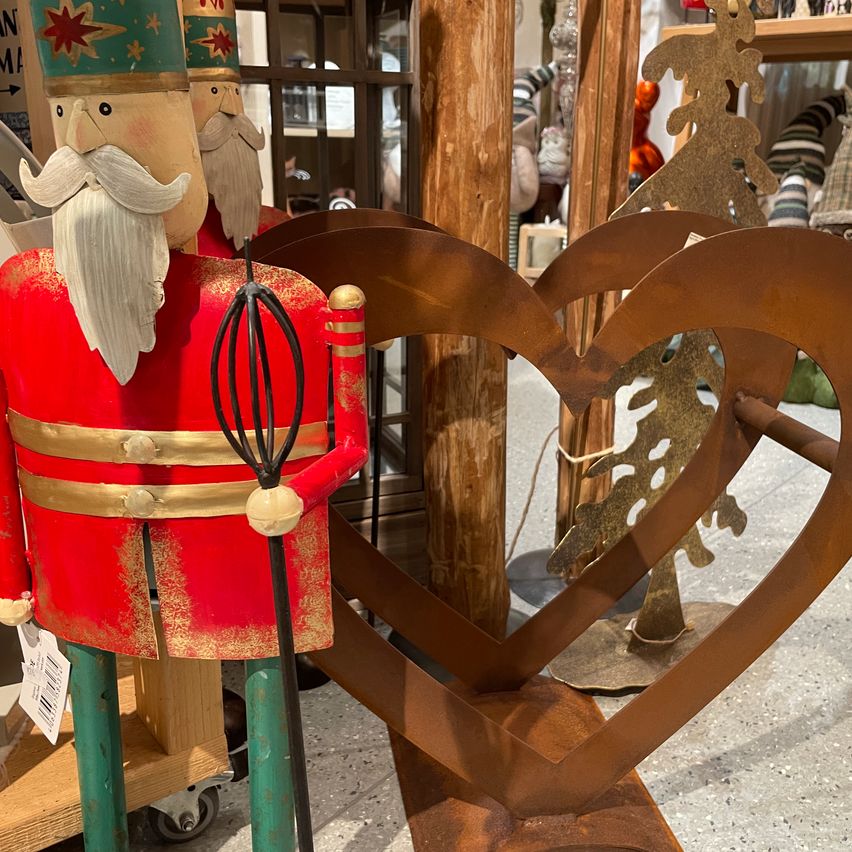 Eine rote Nussknackerfigur mit weißem Bart und grünem Hut steht neben einer herzförmigen Metalldekoration in einem Geschäft.