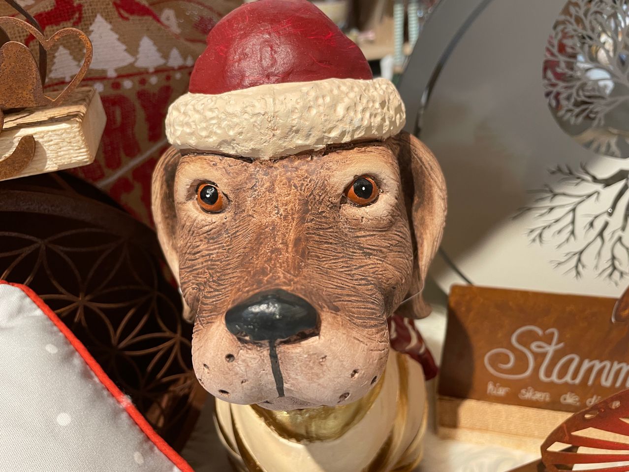 Eine Keramik-Hundefigur mit braunem Fell, die einen Weihnachtsmannhut und einen roten Schal trägt. Der Hund hat braune Augen und eine schwarze Nase. Der Hintergrund besteht aus Weihnachtsdekorationen.