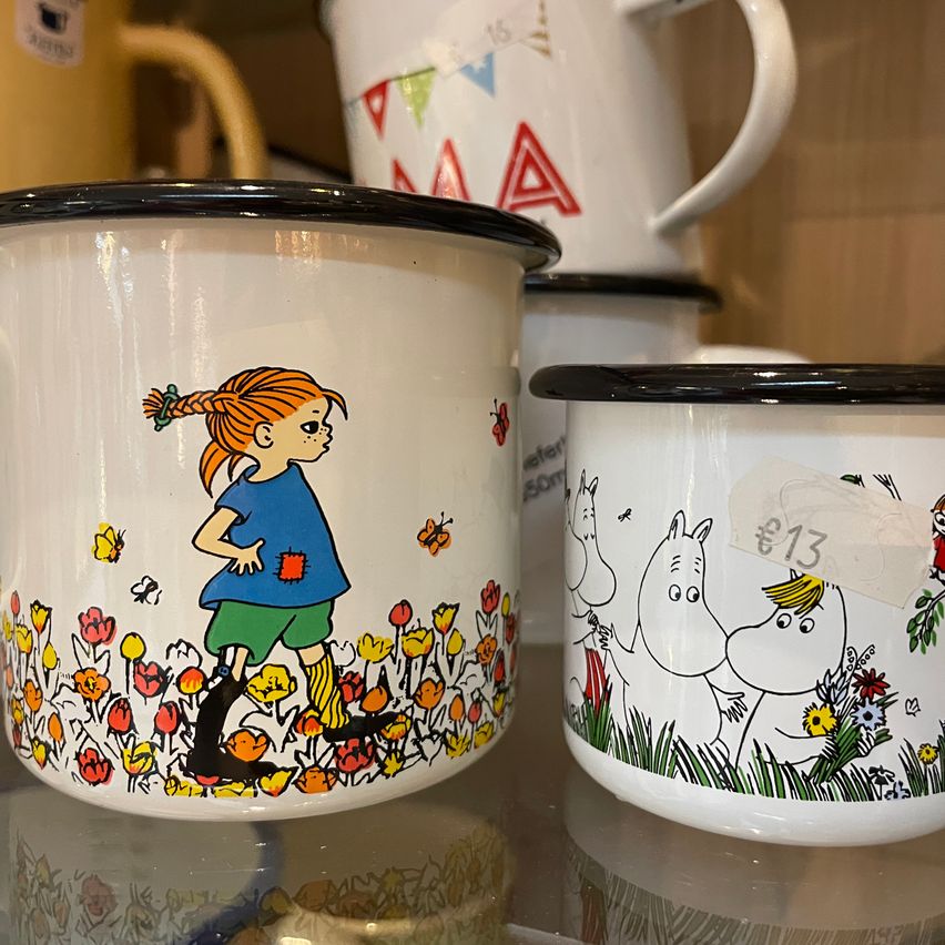 Two white enamel mugs with black rims on display. One has a girl in a field with flowers, and the other has Moomin characters. Price tag on the right mug reads €13.