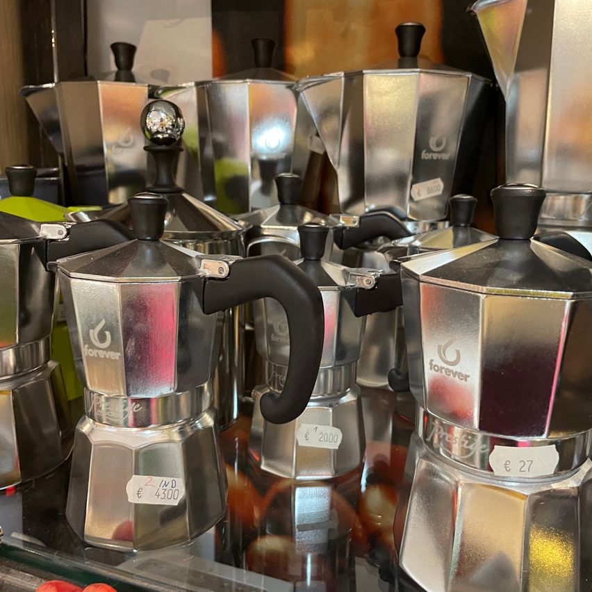 A display of several shiny stainless steel coffee makers with black handles and silver bodies. The foreground features a Forever brand coffee maker priced at 4,300 euros, while a Prestige brand coffee maker priced at 27 euros is visible in the background.