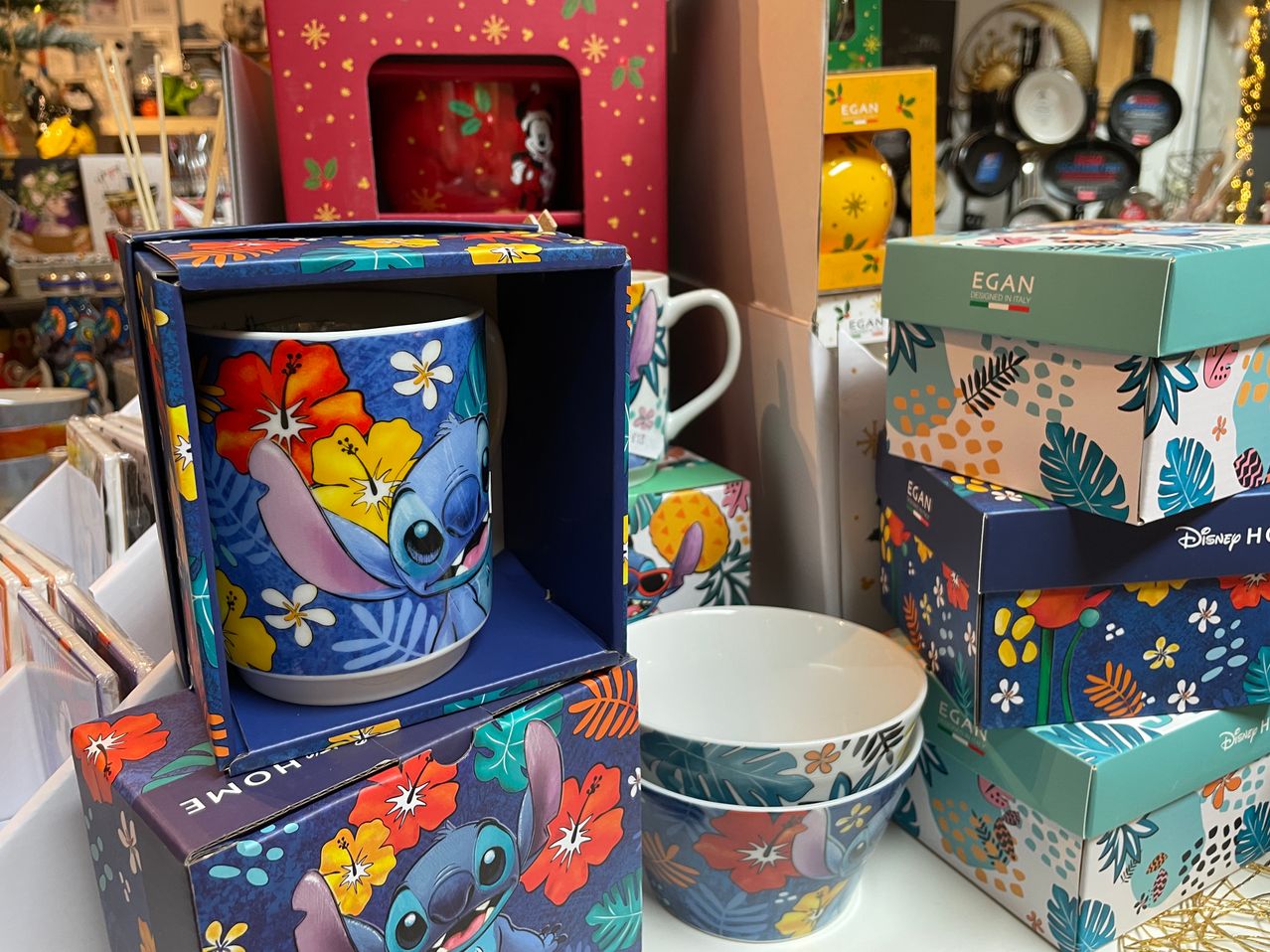 A blue box with a floral design displays a blue mug featuring Stitch. Various colorful boxes and mugs are stacked behind it.