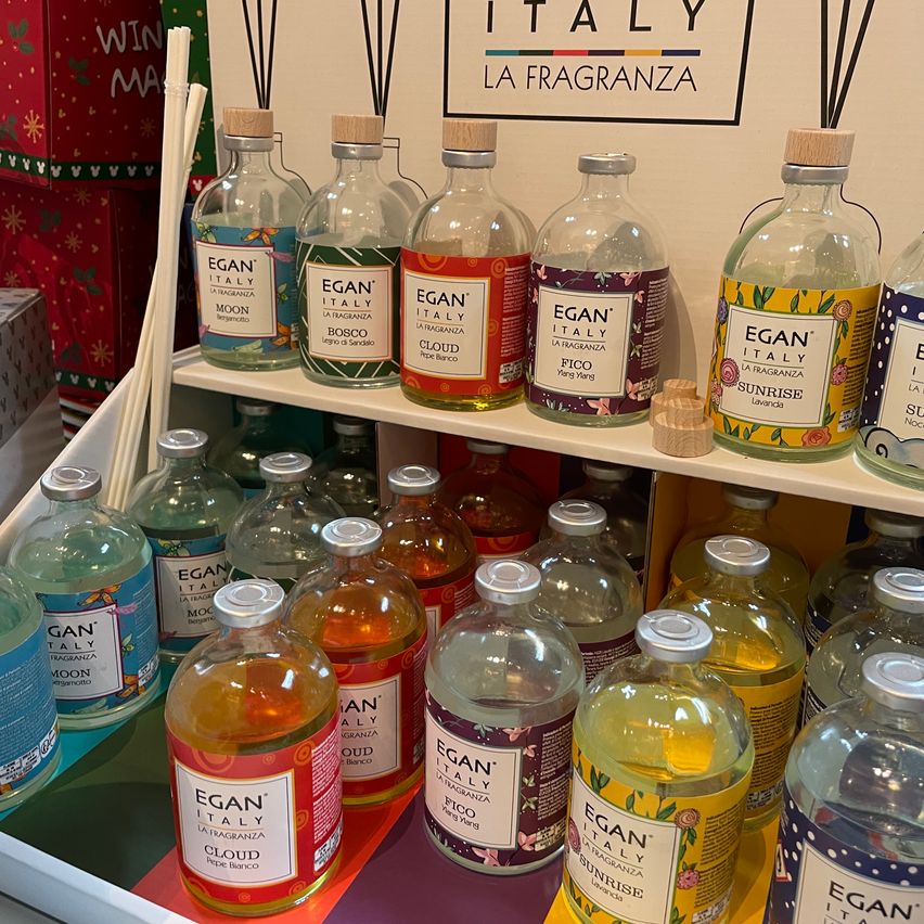 A display of various Egan's Italy scented oils on a white shelf with a red and green background. The bottles are labeled with names like Moon, Bosco, Cloud, and Fico.