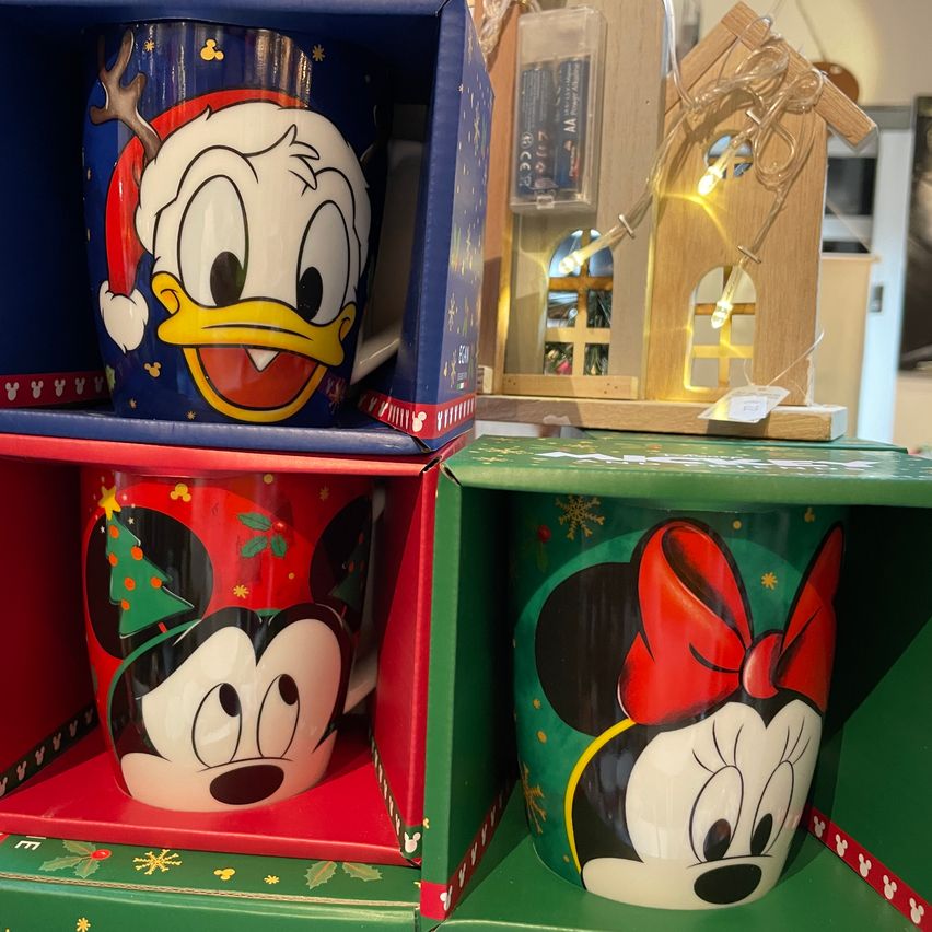 Three themed mugs in gift boxes, one with Donald Duck, another with Minnie Mouse, and the last with Mickey Mouse. A small house-like decoration with lights in the background.