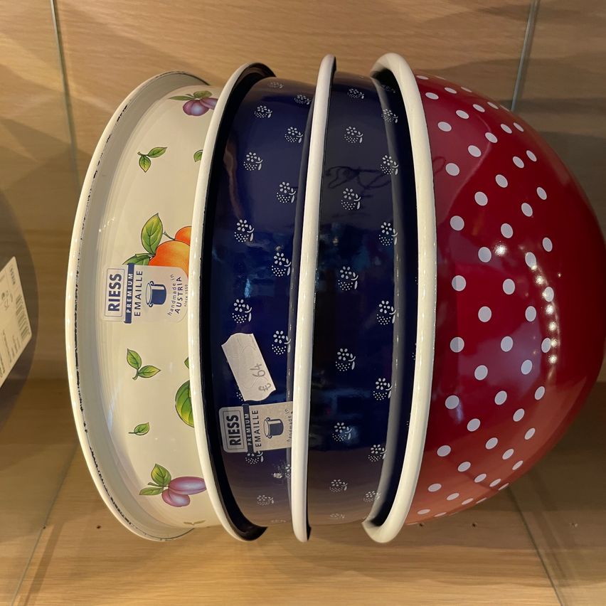 Drei Emaille-Schüsseln, die auf einer Holzoberfläche gestapelt sind. Die Schüsseln haben verschiedene Designs: eine weiße mit Früchten, eine blaue mit Blumenmustern und eine rote mit weißen Punkten.