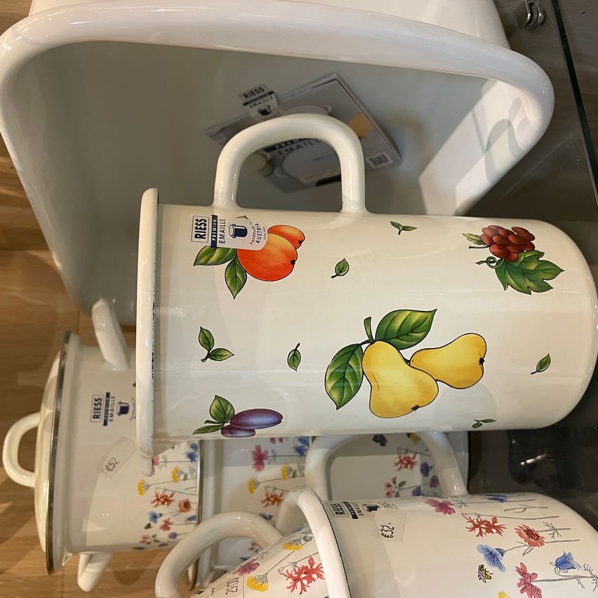 Ein Stapel weißer Keramikbecher mit Fruchtdesigns auf einer Holzoberfläche. Der oberste Becher zeigt Orangen, Birnen und Pflaumen. Darunter ein Becher mit Blumendesigns.